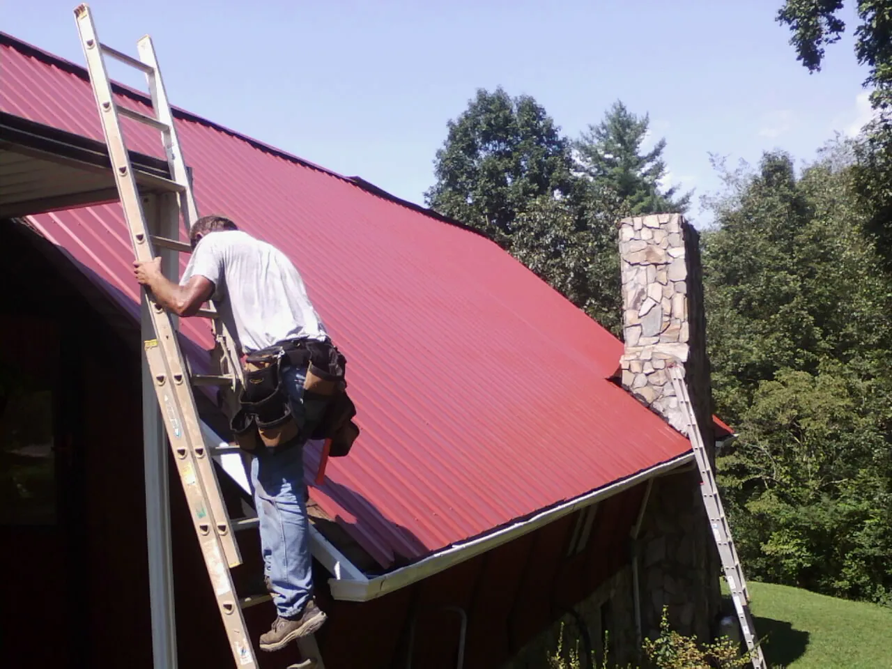 Roofer on a ladder beside a red metal roof and stone chimney — Soffit & Fascia Repair by Roofing Brothers in Kilgore